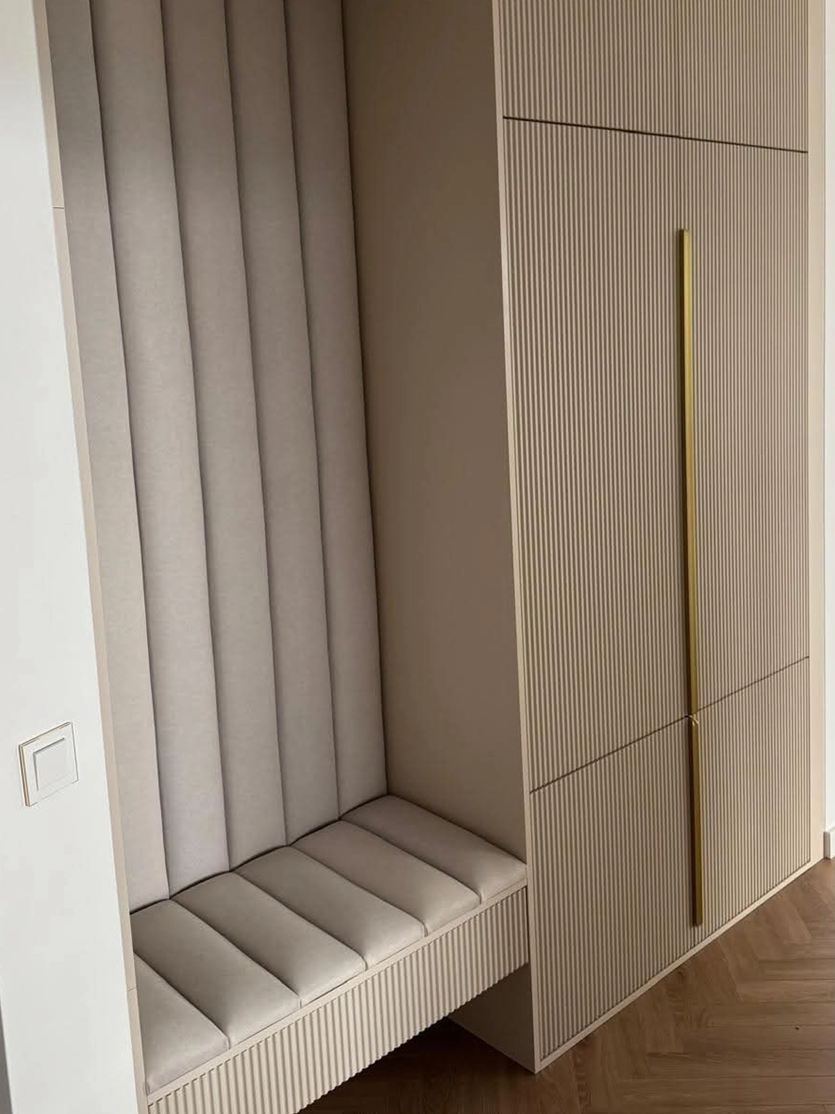 Garderobe nach Maß im Flur: offene Sitznische mit gepolsterten Wandpaneelen und Sitzpolster in Beige, daneben eine deckenhohe Schrankfront mit feiner Rillenstruktur und langer goldener Griffleiste.