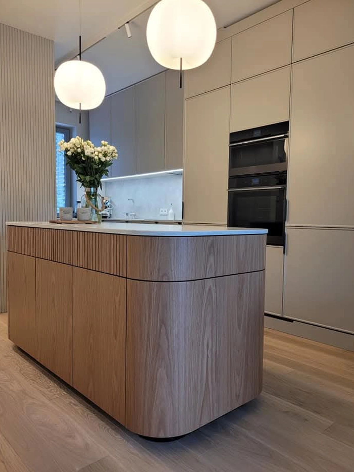 Moderne Einbauküche nach Maß mit grifflosen Fronten in Creme: eine Kücheninsel mit abgerundeten Ecken, Lamellenfront in Holzoptik und heller Steinarbeitsplatte, dahinter ein Hochschrank mit zwei Einbaubacköfen.