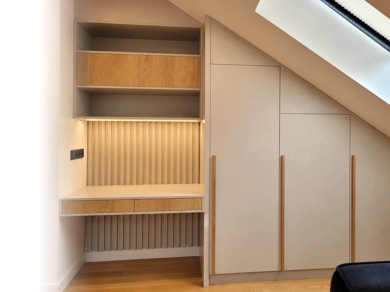 Einbauschrank nach Maß unter einer Dachschräge: raumhohe Fronten in mattem Greige mit schmalen Eichengriffen, daneben eine Nische mit offenen Regalen, Schreibtischplatte und Schubladen in Eiche.